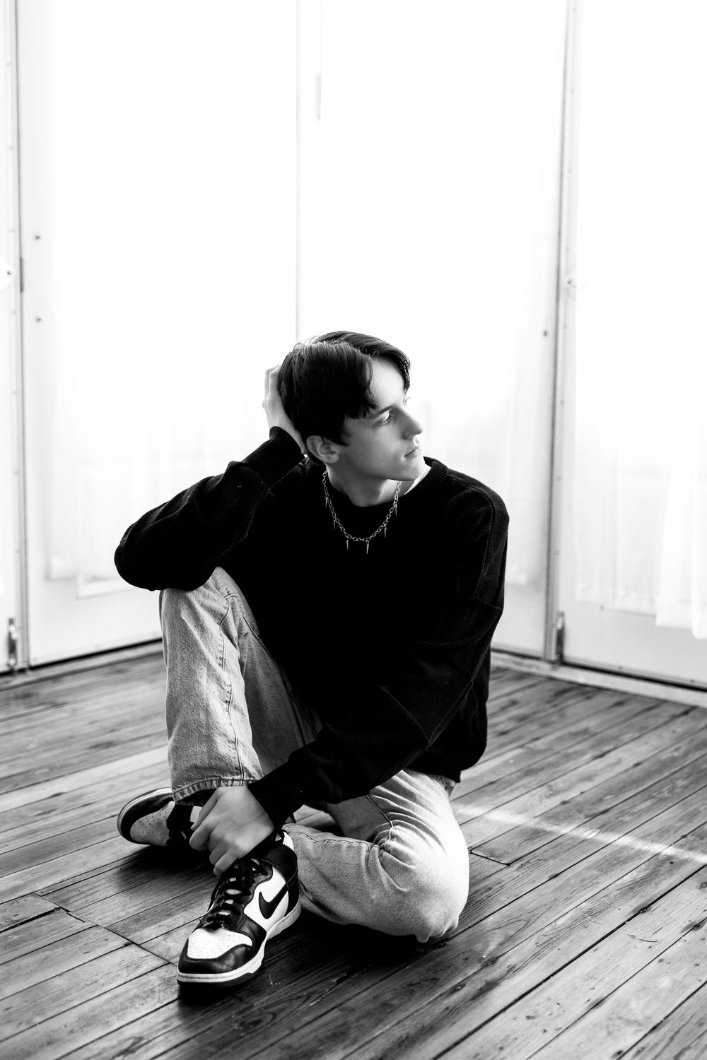Black and white photo of young man in dark sweater sitting on wooden floor looking to the side.