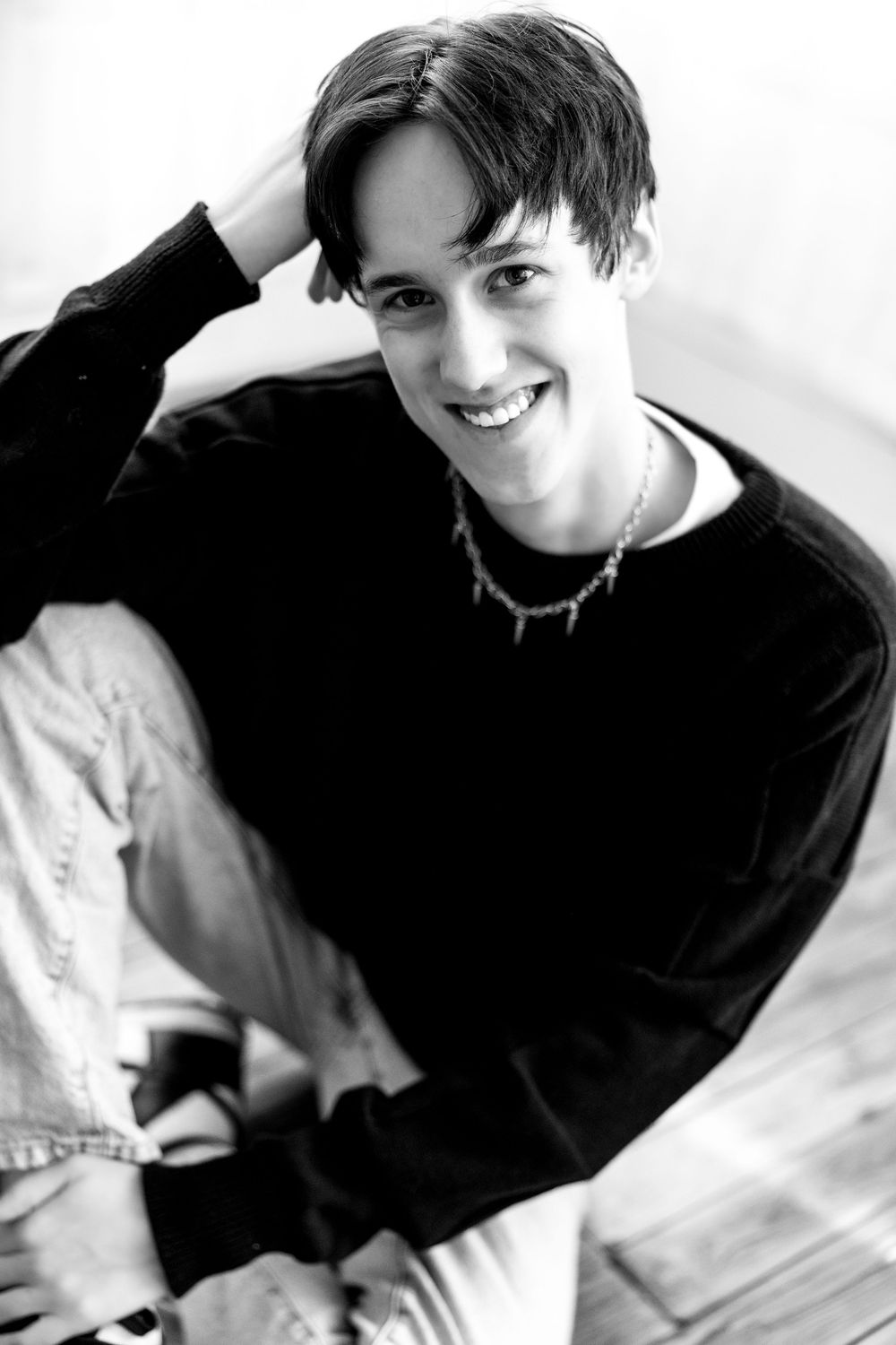 Black and white overhead shot of smiling young man in black sweater with chain necklace on floor.