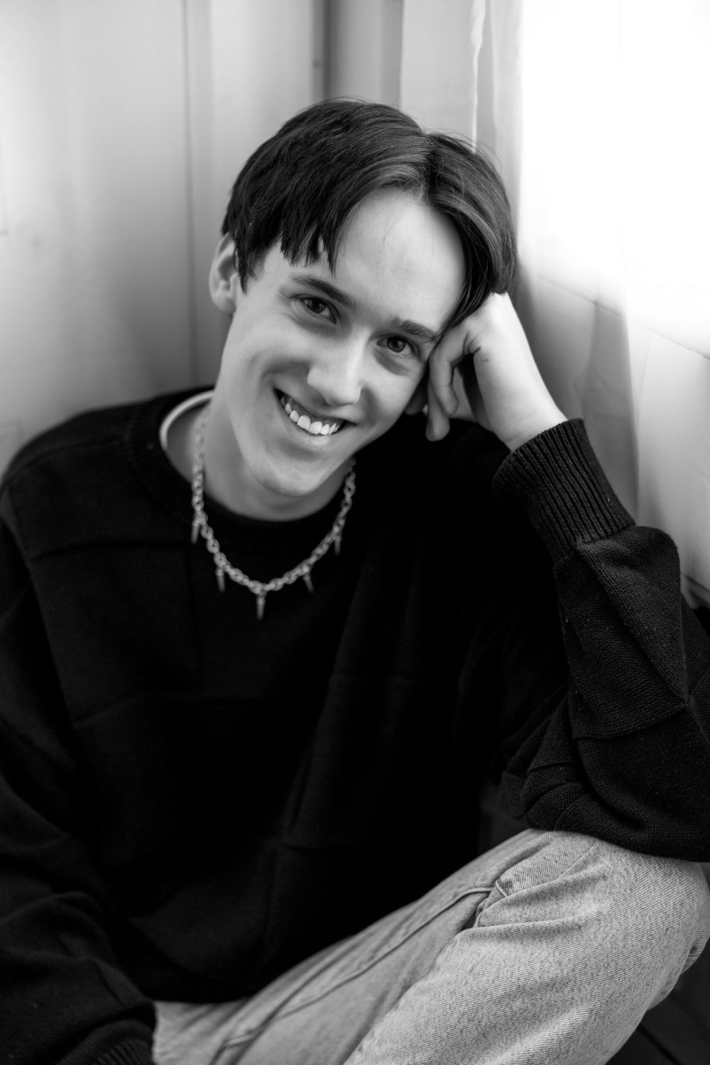 Smiling young man in black sweatshirt and chain necklace posing casually in a black and white portrait photo.