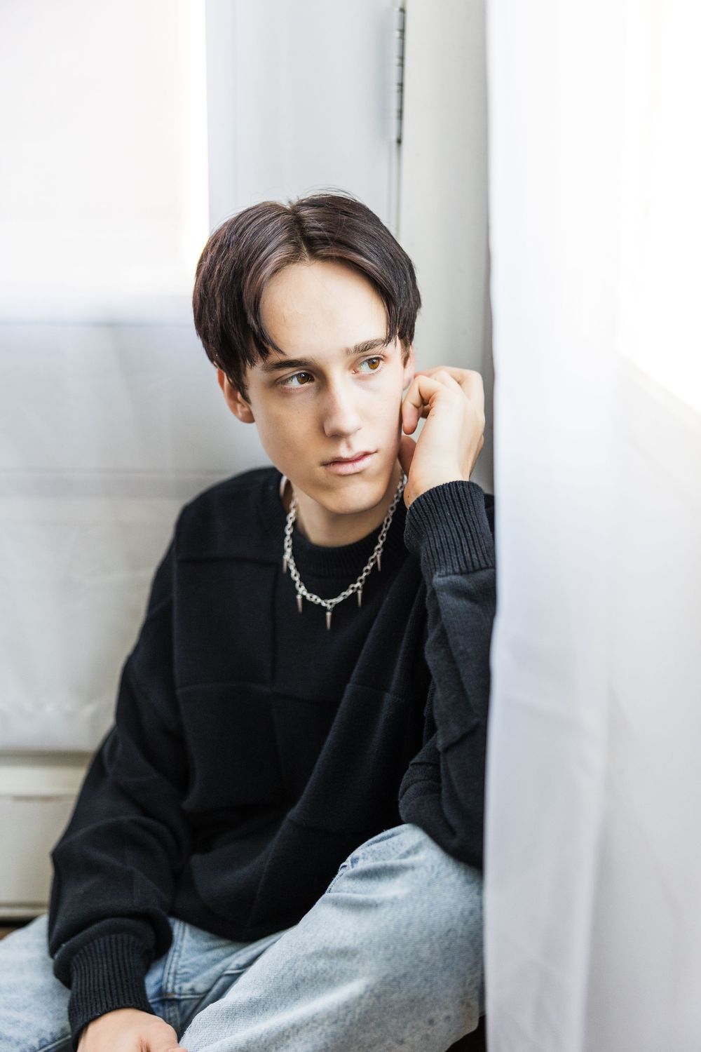 Young man in black sweatshirt with chain necklace sitting against a white wall with pensive look.