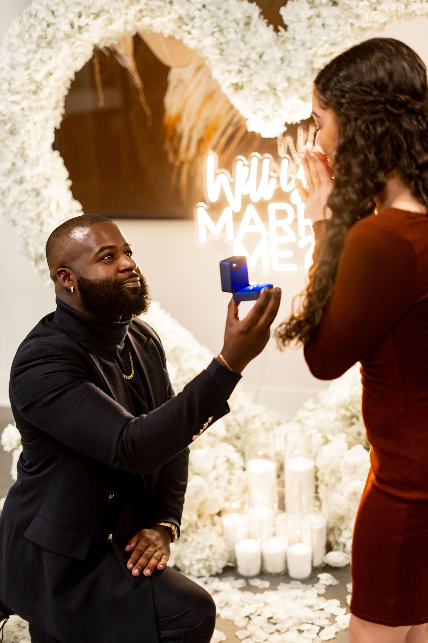 A romantic proposal moment with neon lights and candles in the background while presenting a ring box.