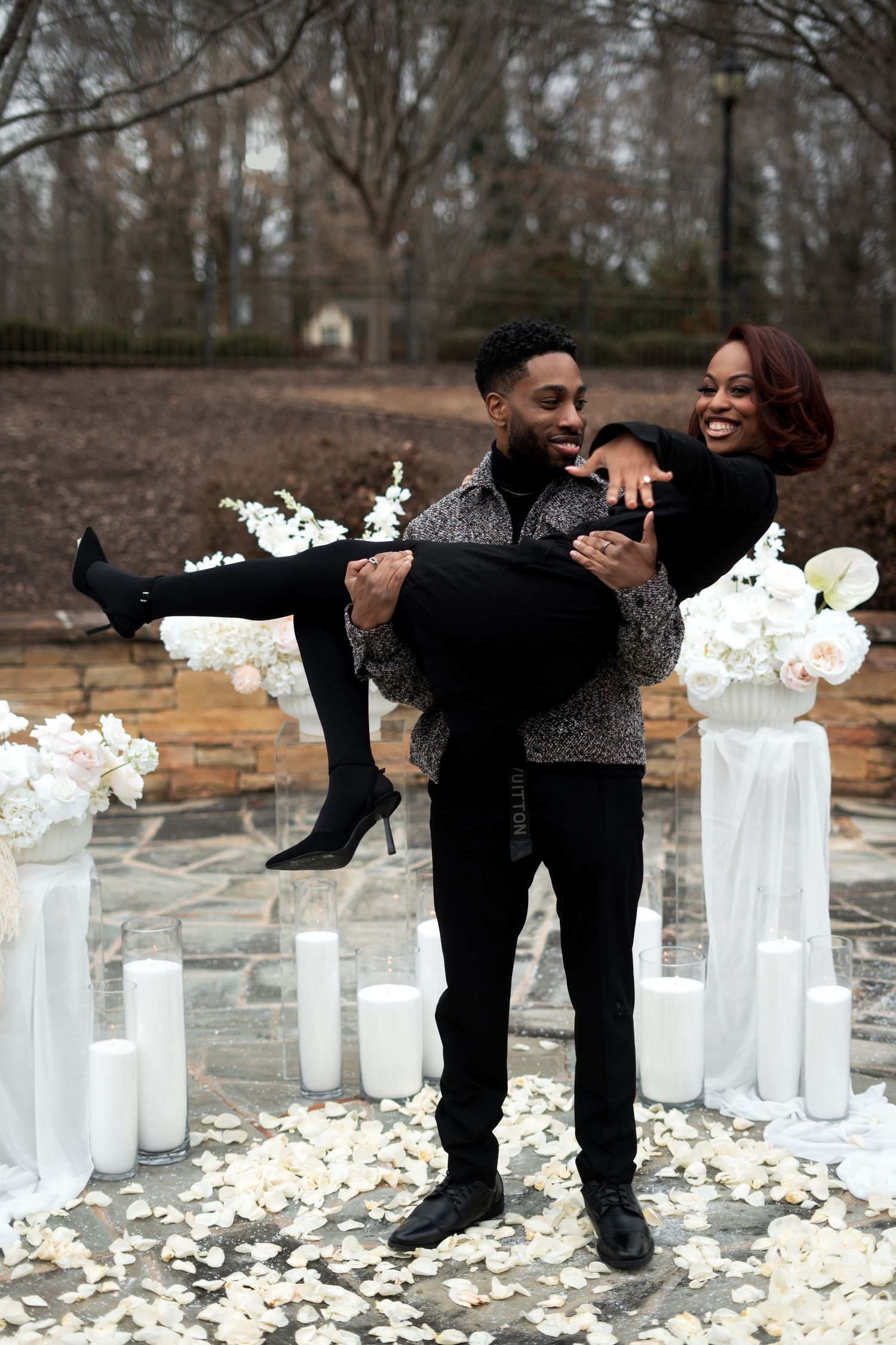 Couple sharing a joyful moment during a romantic outdoor proposal setup with white candles and rose petals on the ground.