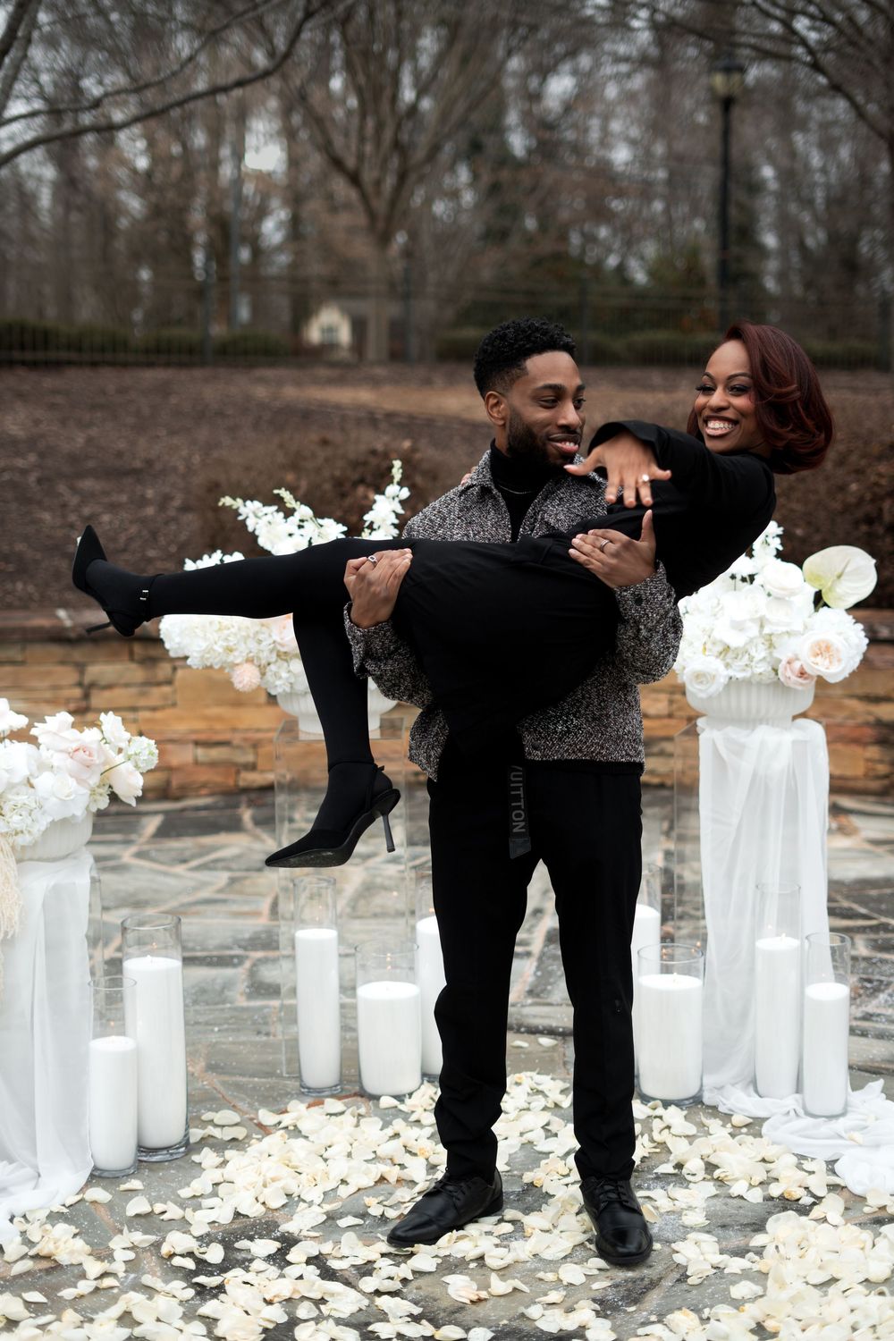 Couple sharing a joyful moment during a romantic outdoor proposal setup with white candles and rose petals on the ground.