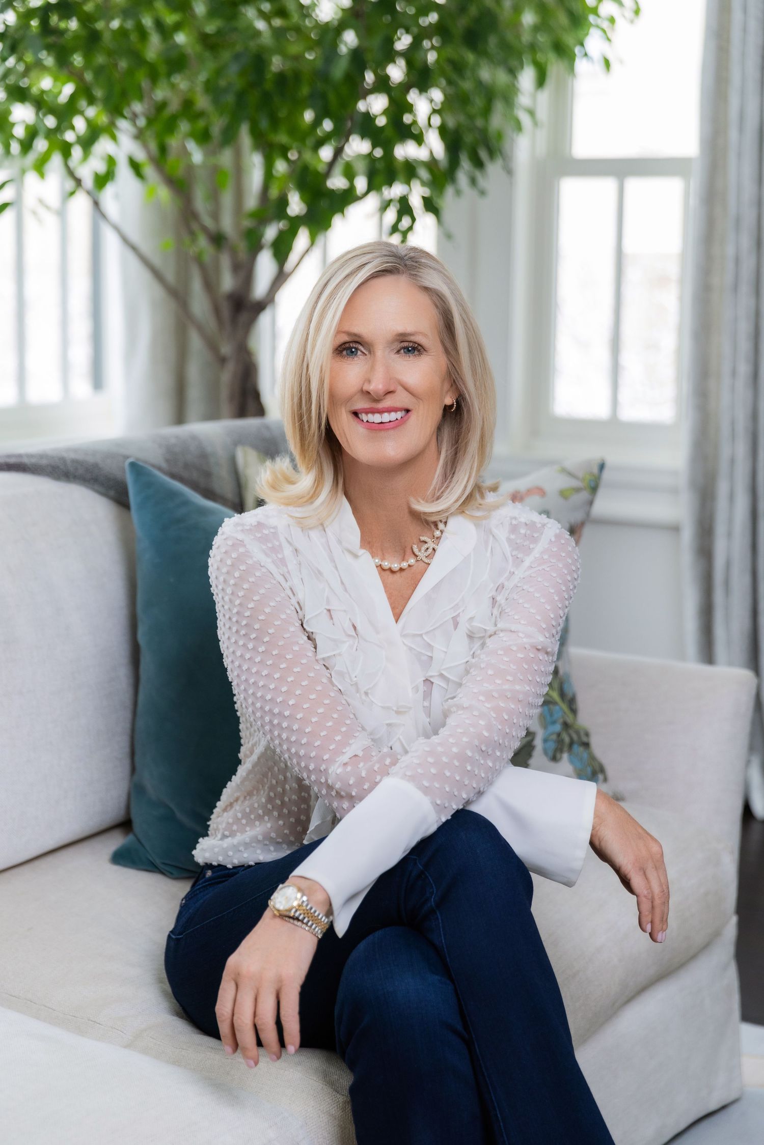 Professional portrait of someone seated on a grey sofa wearing a white textured sweater and dark pants with a houseplant behind.