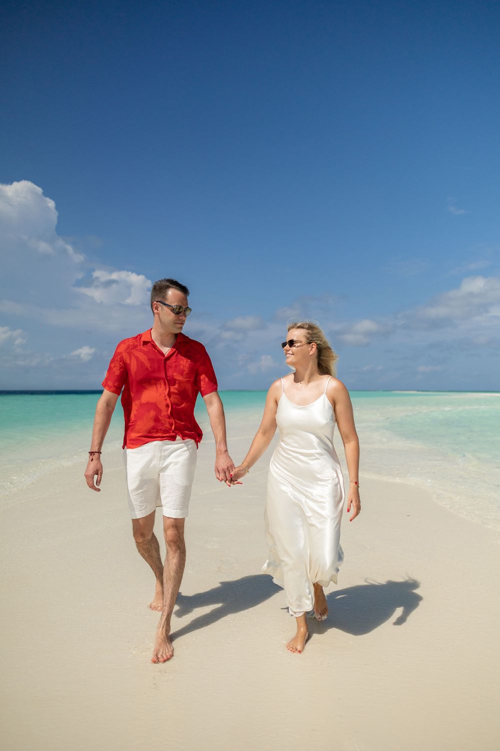 A couple walks hand in hand on a pristine white sand beach with turquoise waters and blue sky in the Maldives.