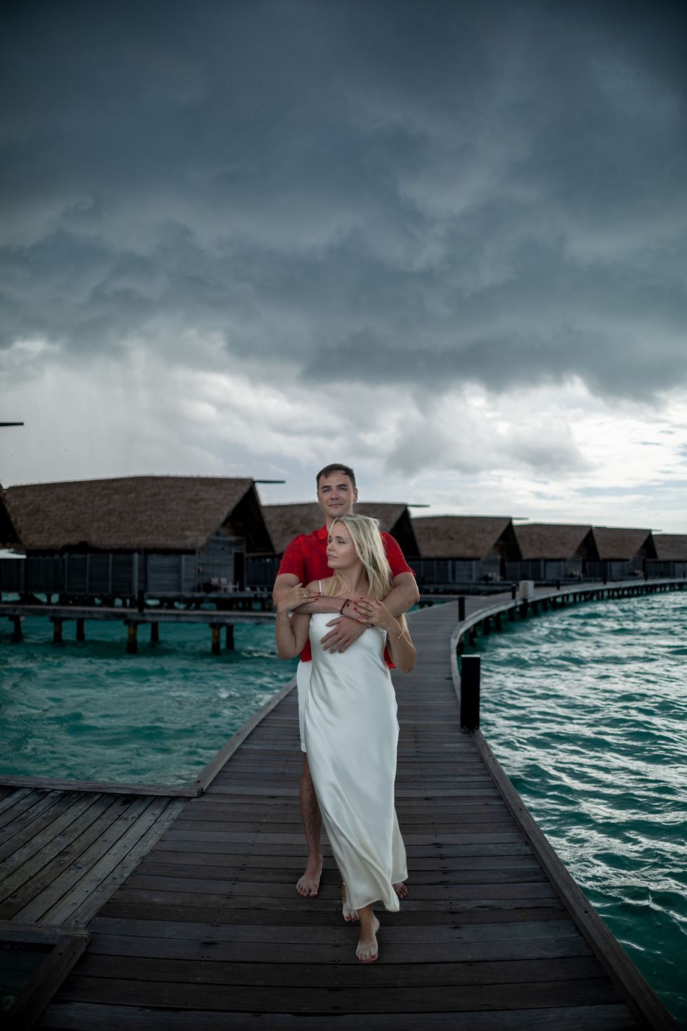 A couple embraces on a wooden walkway over turquoise waters at a Maldives resort under dramatic stormy skies.