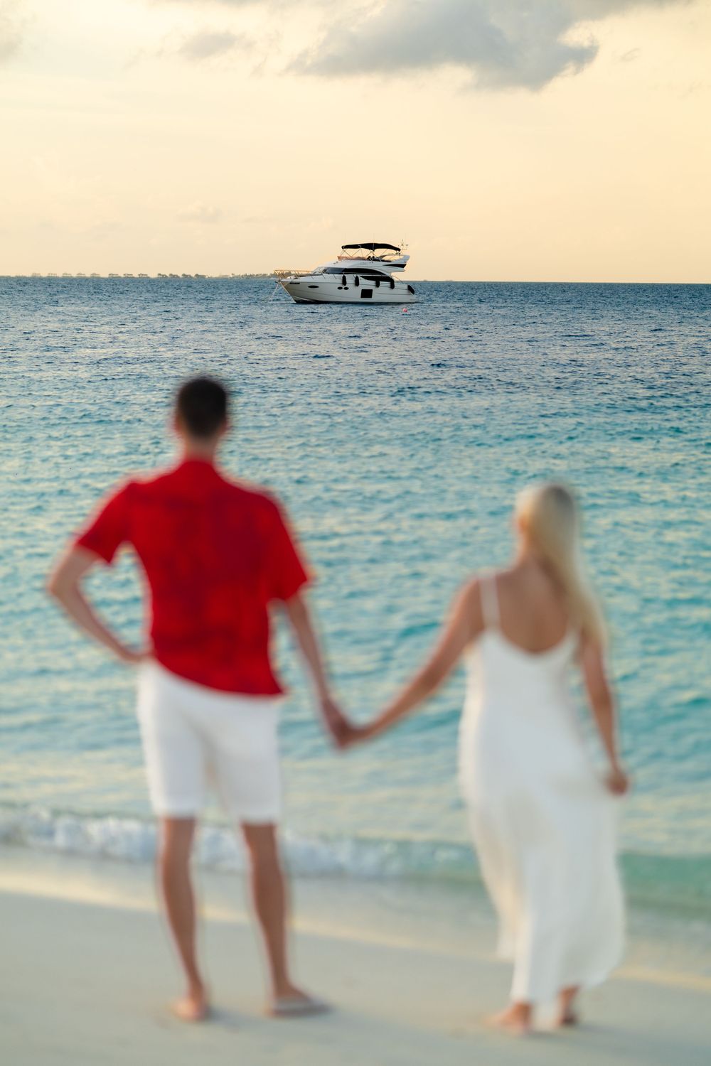 A couple in red and white outfits walking hand in hand along a pristine beach with turquoise waters and a boat in distance.