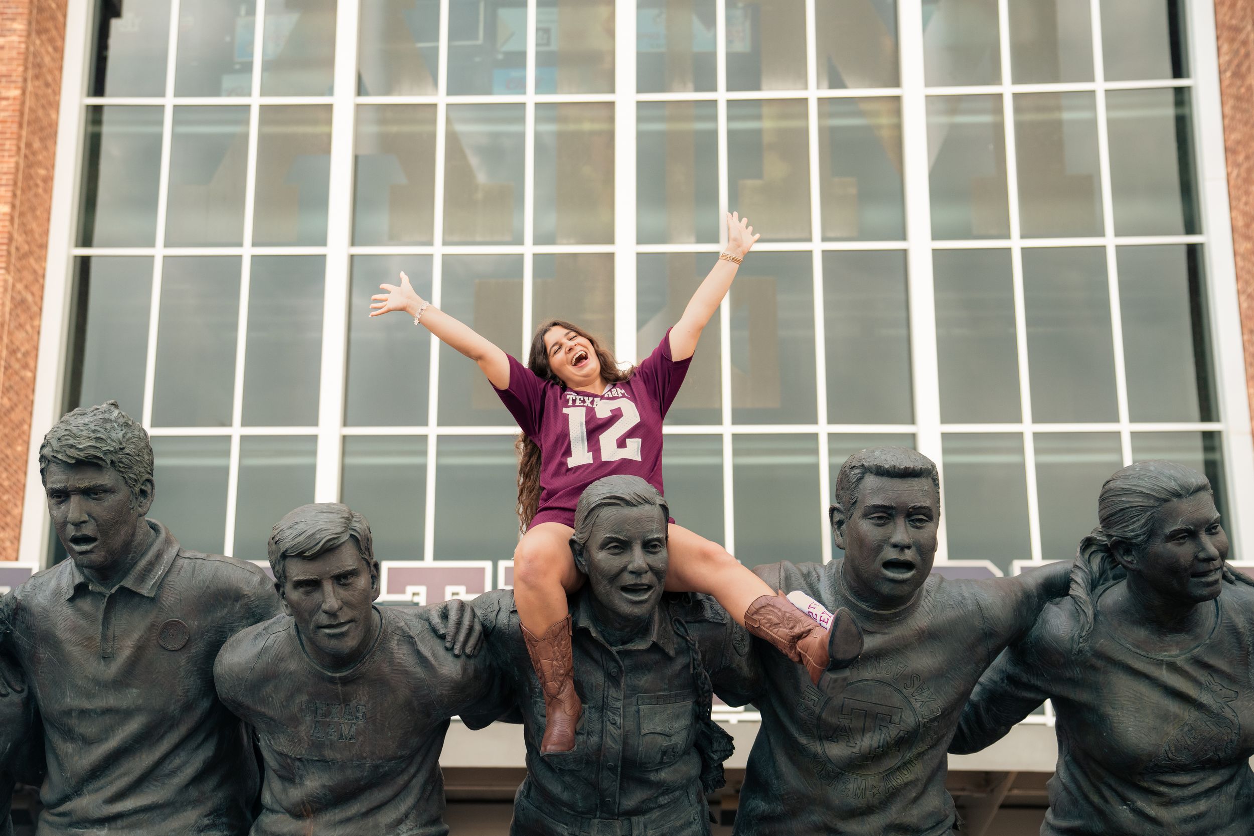 Student celebrates on sports statues outside athletic facility.