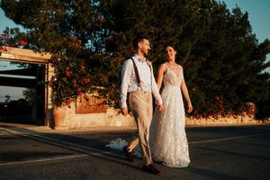 A couple in wedding attire walks together on a sunset-lit street with trees in the background.