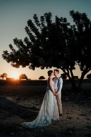 A couple shares an intimate moment at sunset beneath a tree silhouette in a rustic outdoor setting.