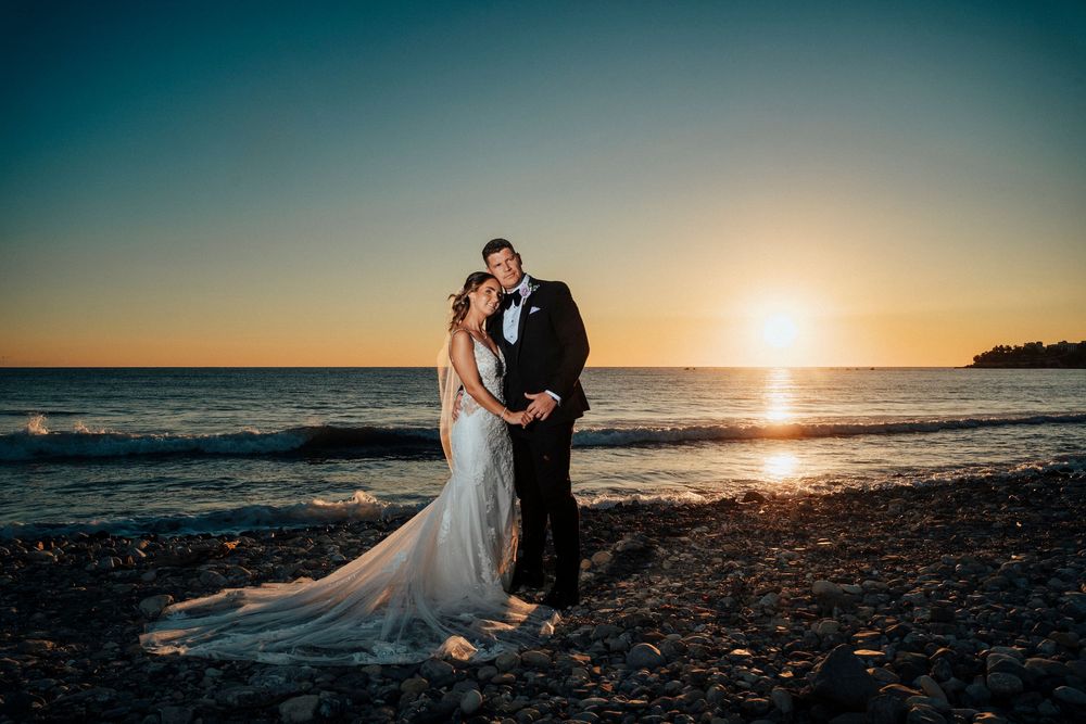 A couple in wedding attire embraces on a rocky beach at sunset with golden light reflecting off the ocean.