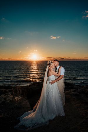 Silhouettes of a couple in wedding attire embracing on a rocky beach at sunset with dramatic ocean views.