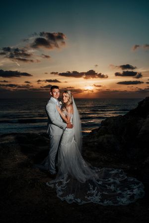 A romantic wedding couple poses against a dramatic ocean sunset backdrop on rocky coastal terrain.