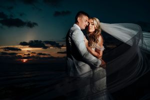 Newlywed couple embraces during sunset beach wedding with flowing veil creating romantic silhouette against orange sky.