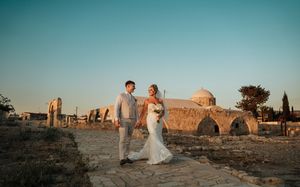 A couple poses in a golden desert landscape during sunset with ancient ruins and cypress trees in the background.
