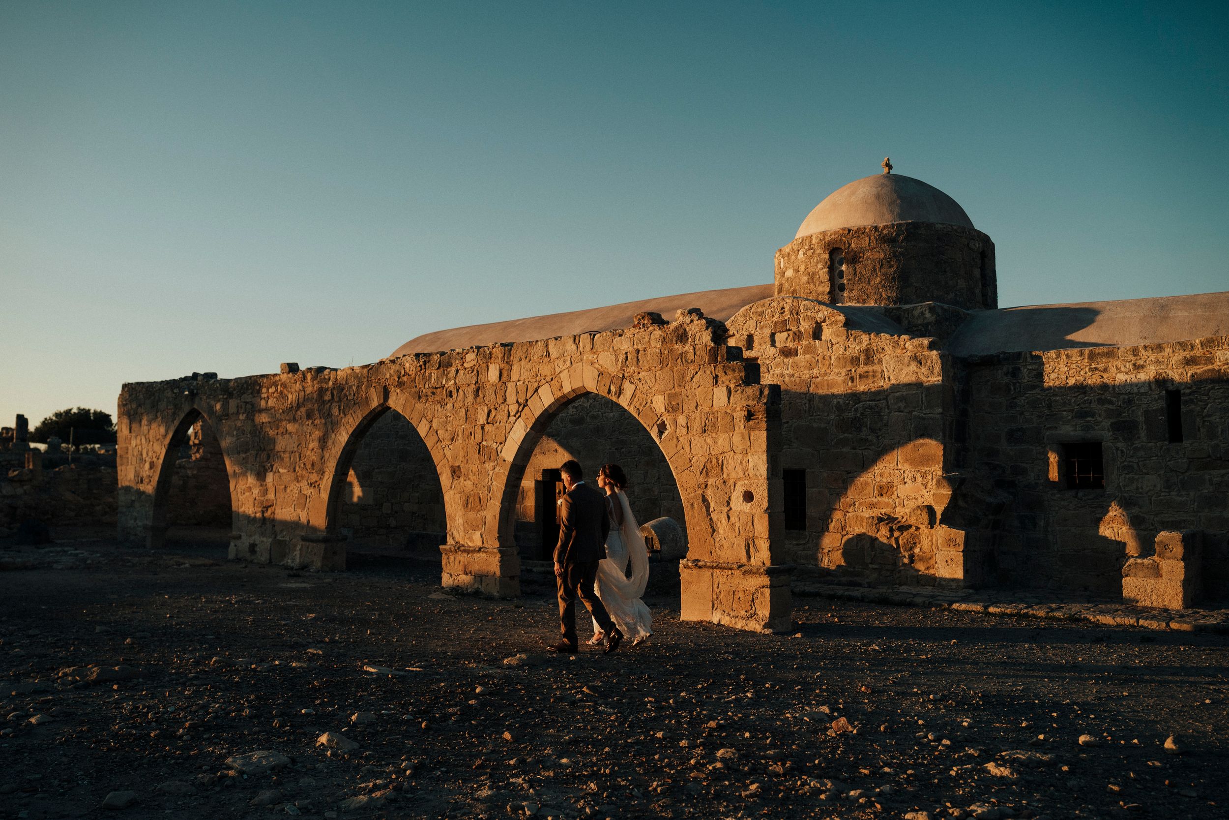 Ancient stone ruins and archways cast long shadows in warm sunset light across a desert landscape.