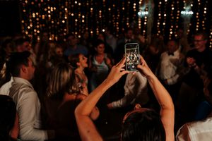People raise their phones to capture a moment at a dimly lit party with string lights in the background.