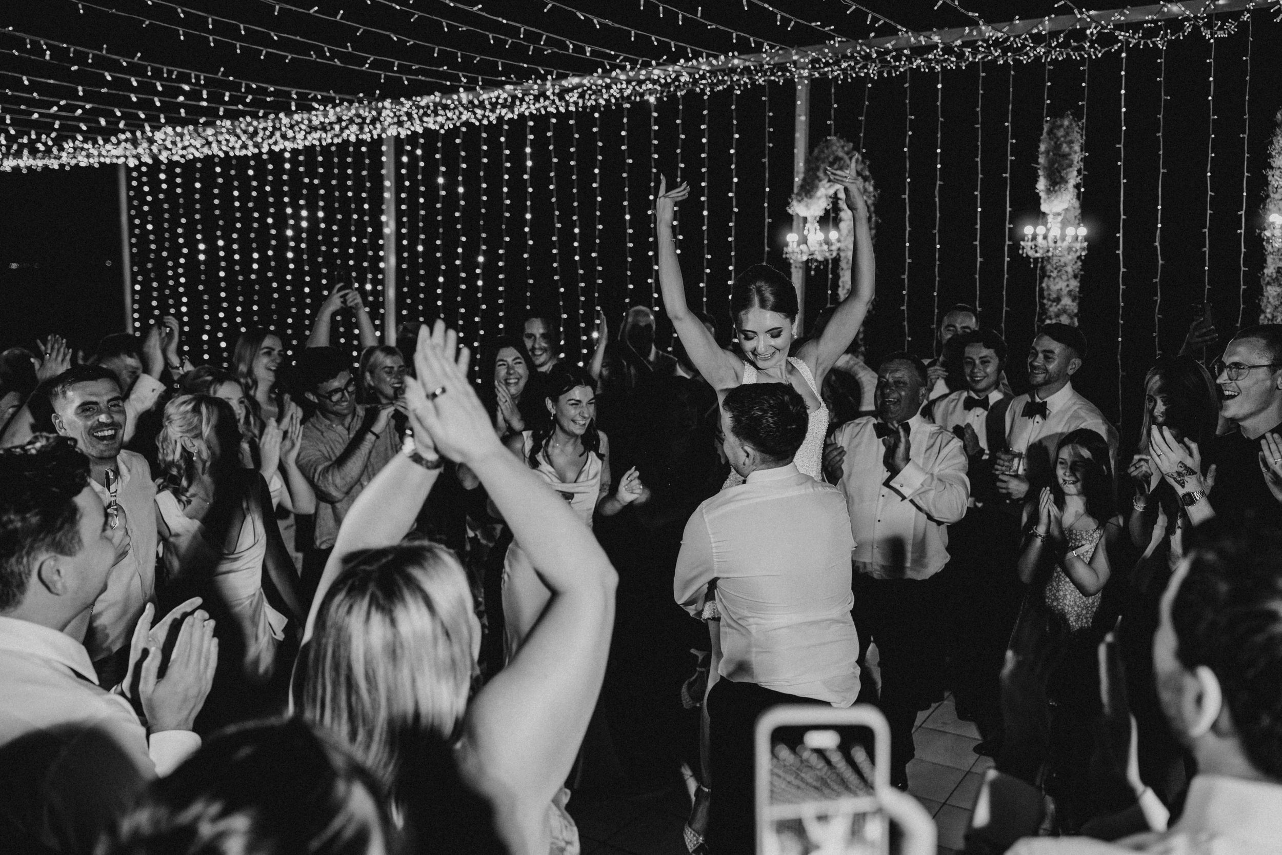 People dancing and celebrating at a wedding reception under glittering string lights and chandeliers.