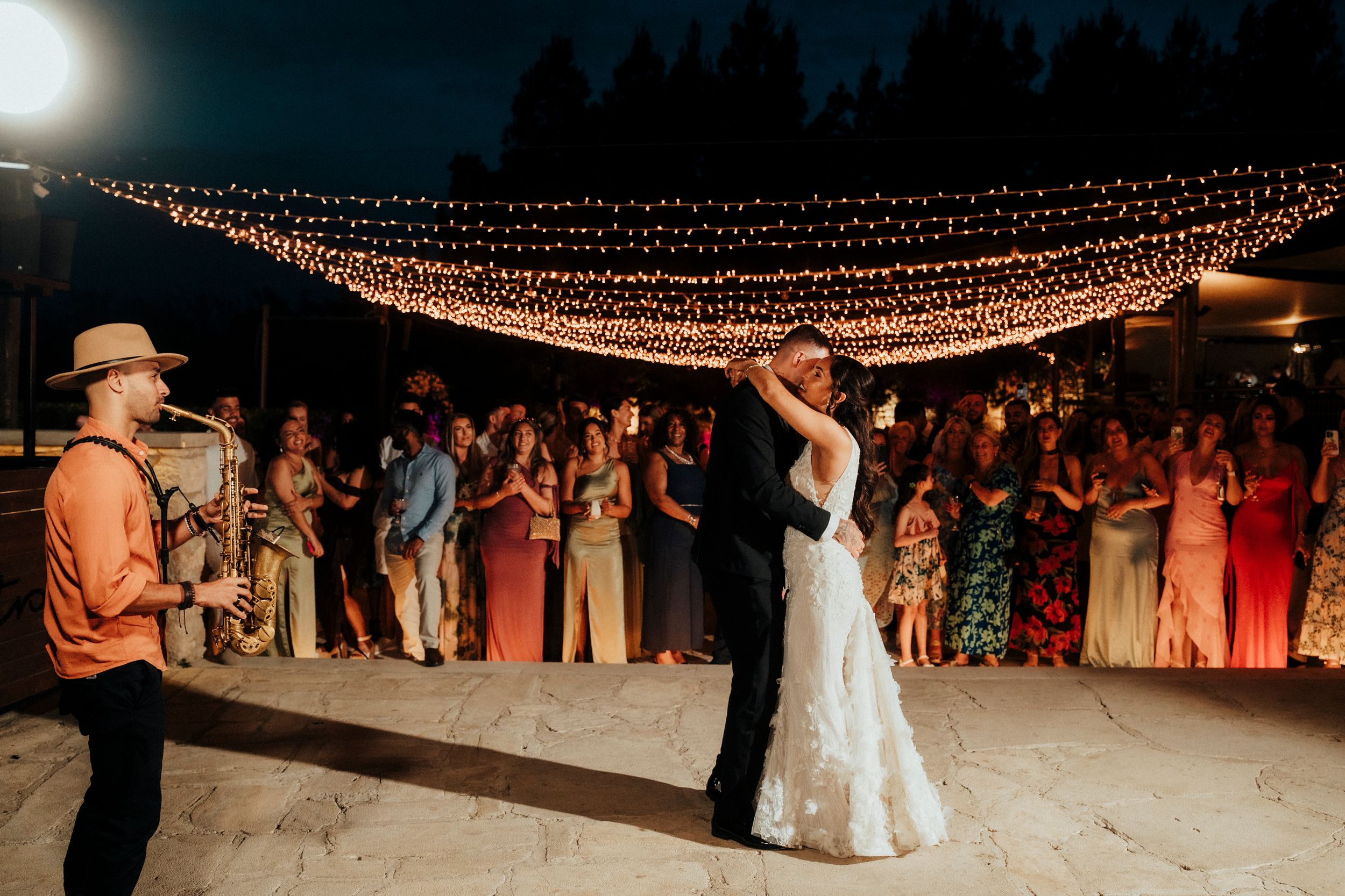 String lights illuminate an outdoor wedding reception dance floor as guests celebrate under the night sky.