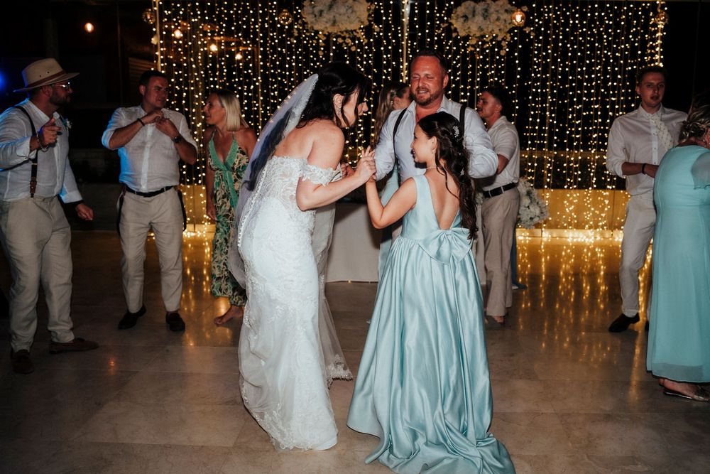 Wedding guests dance and celebrate on a dimly lit dance floor decorated with twinkling string lights in the background.
