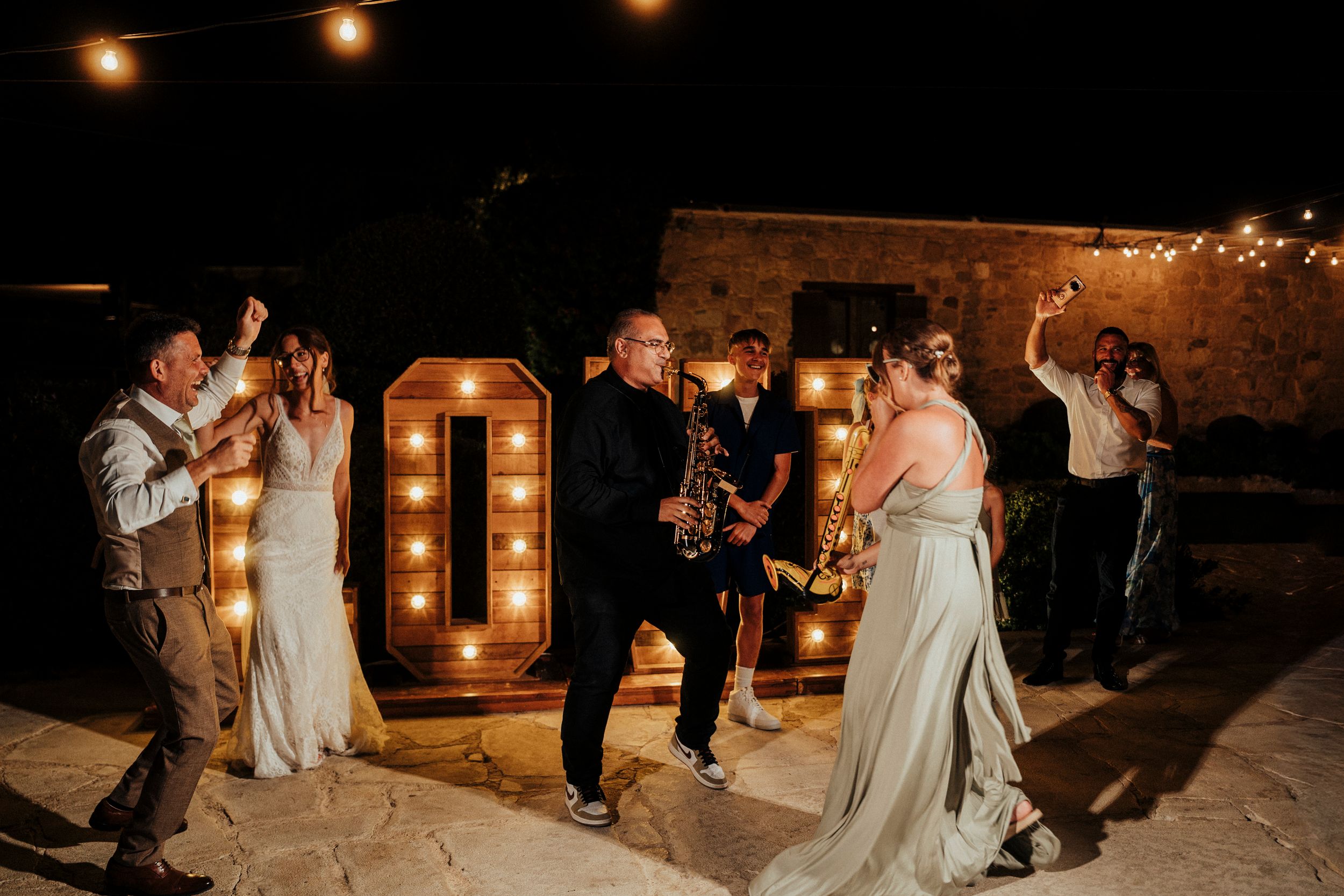 Wedding party dancing and celebrating at a romantic evening reception with warm string lights overhead.
