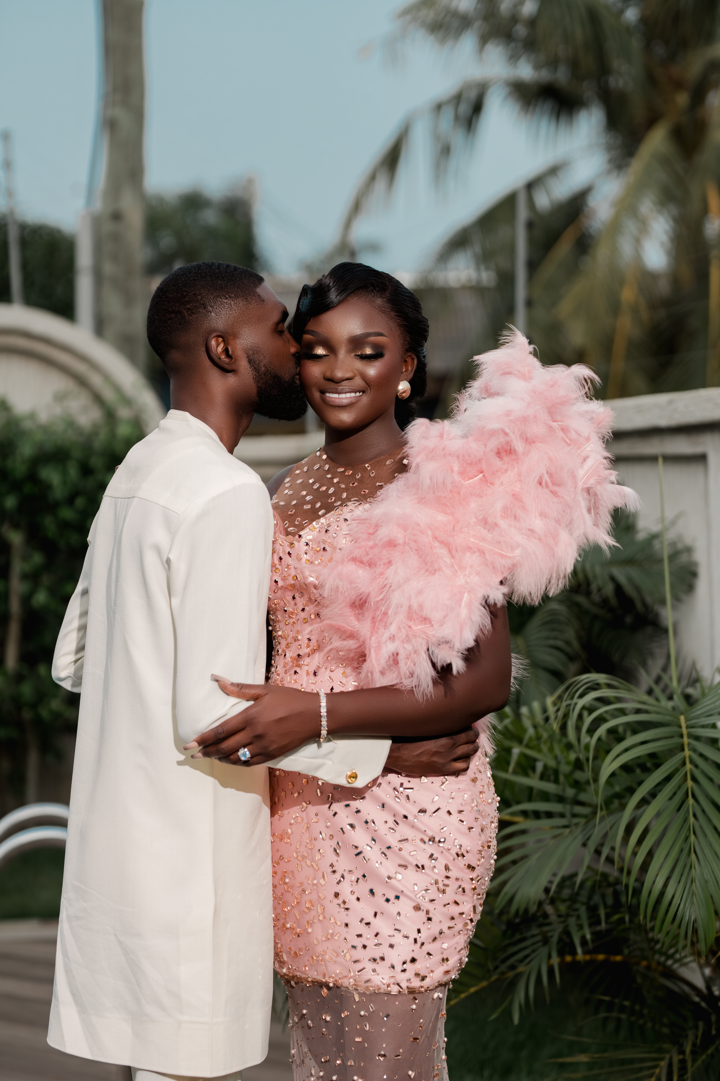 Outdoor portrait of a couple in traditional ivory attire; bride holding a large pink feathered fan.