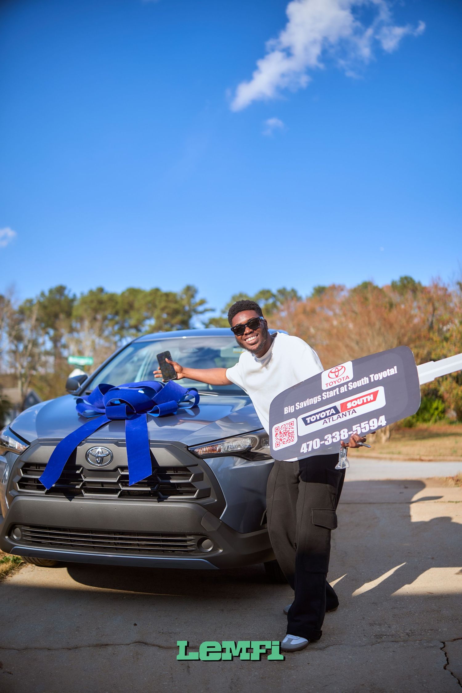 A new car with a large blue bow sits on a sunny road during a vehicle delivery presentation.
