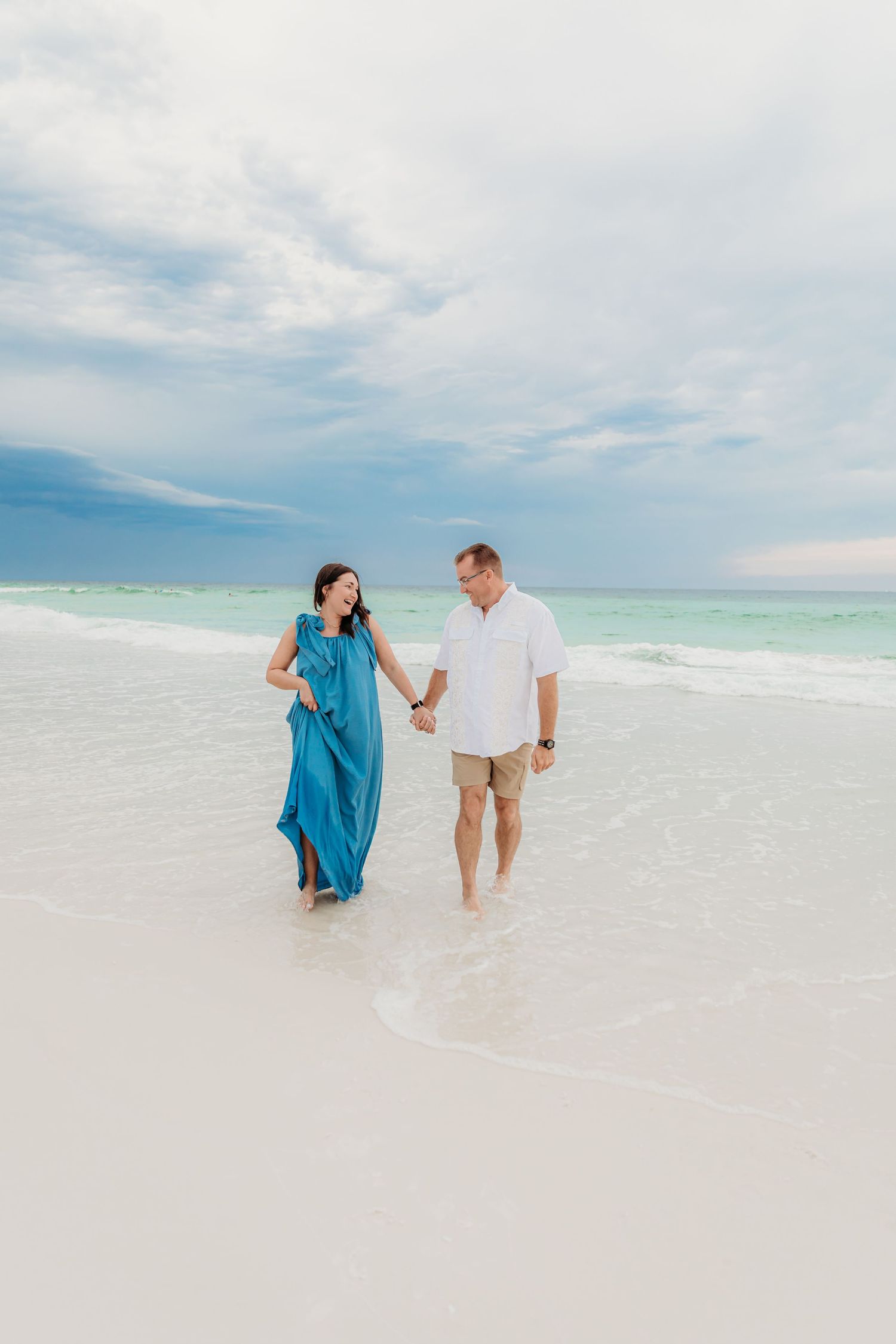 A couple walks hand in hand along a pristine white sand beach with turquoise waters and cloudy skies in the background.