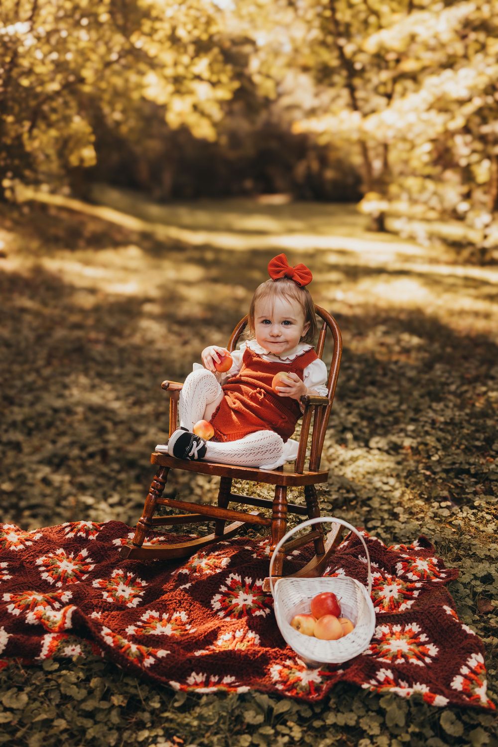 Toddler girl in red dress sitting in rocking chair on floral blanket with fruit basket in autumn park in Oak Ridge, TN.