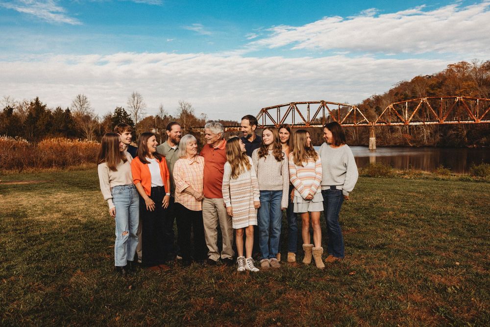 Large family group portrait outdoors near a rustic bridge, posing on green grass with autumn trees in background in Oak Ridge, TN.