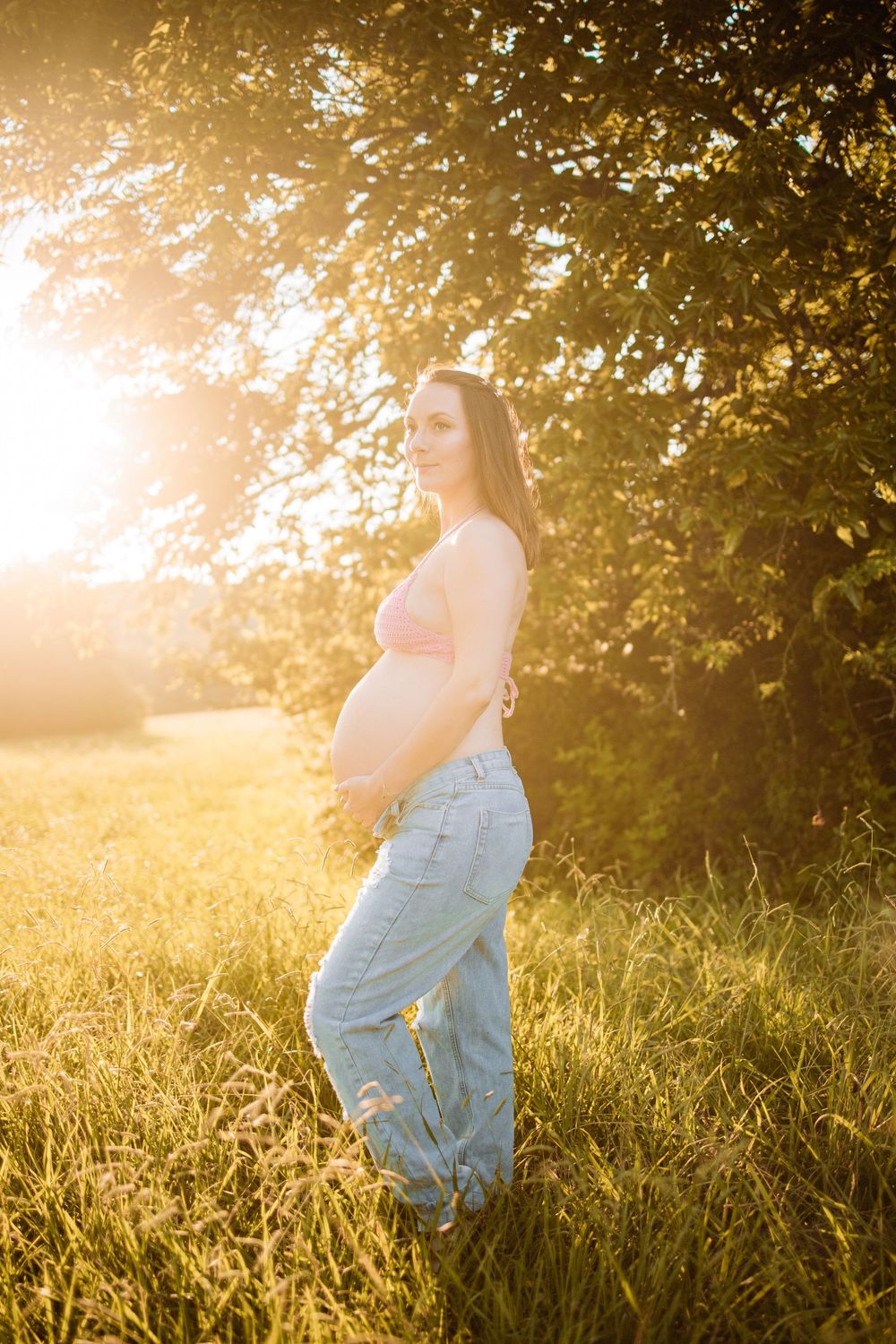 Pregnant woman in pink crop top and jeans standing in sunlit meadow with golden hour light and green trees in Oak Ridge, TN.