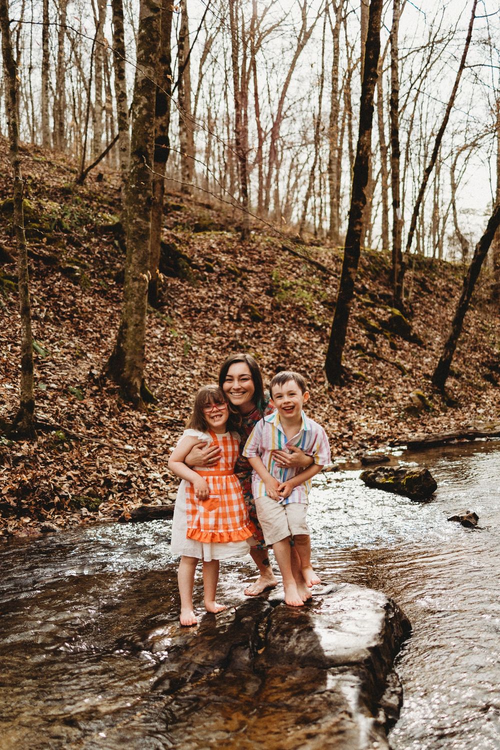 A woman and two young children stand on rocks in a forest creek surrounded by autumn trees and fallen leaves at Norris Dam State Park.