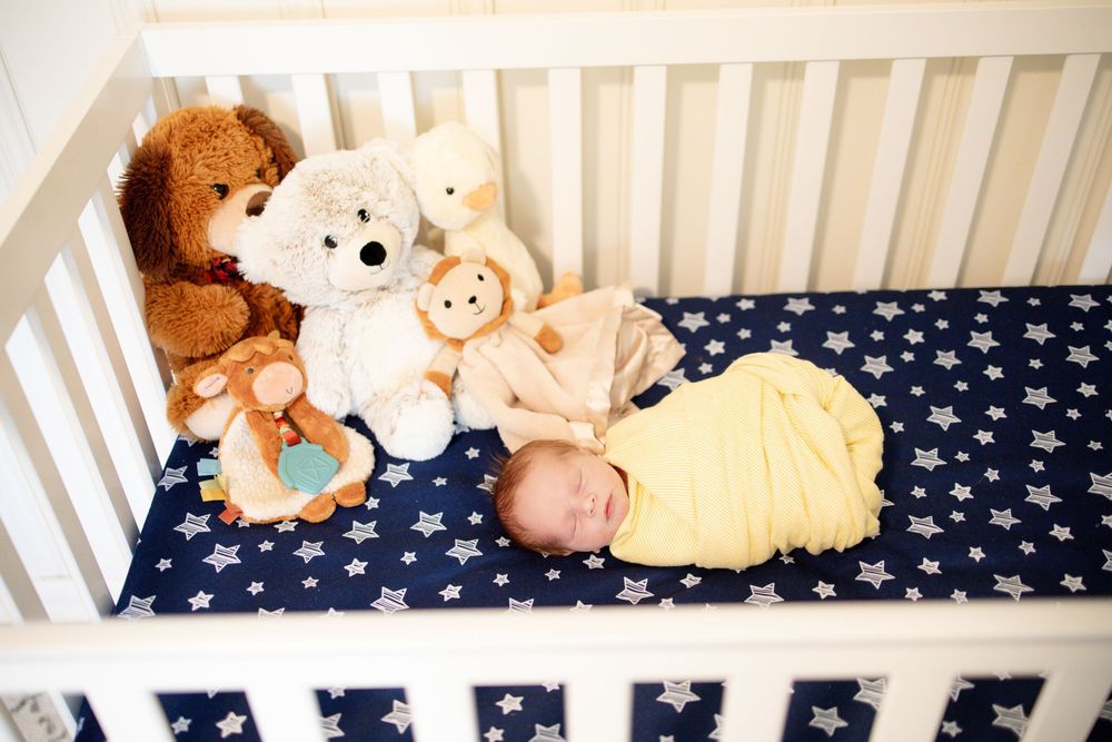 Sleeping newborn baby in yellow swaddle inside white crib with star sheet surrounded by stuffed animals in Knoxville, TN.