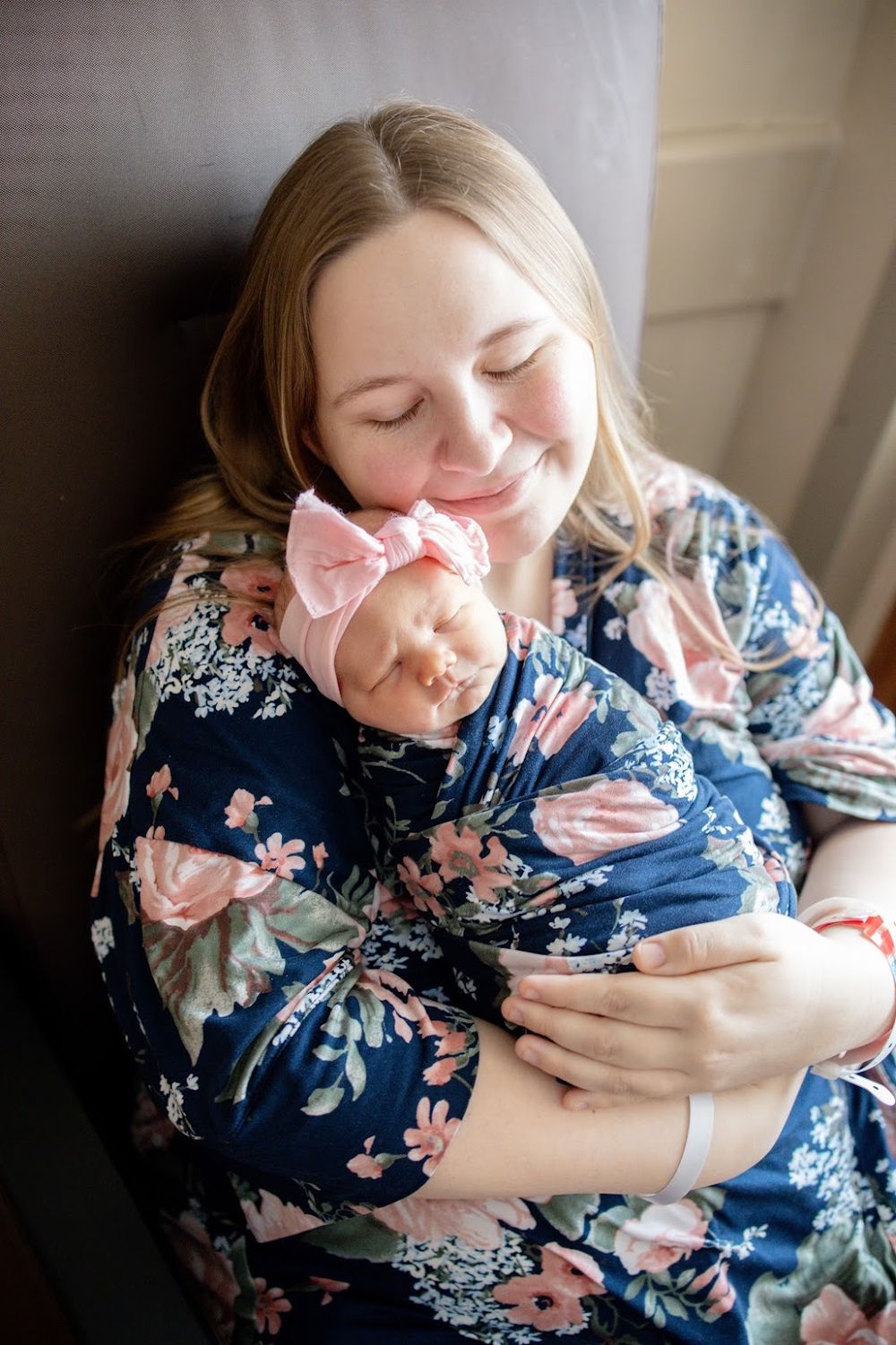 A smiling mother in a floral dress tenderly holds her sleeping newborn baby wearing a pink bow headband.