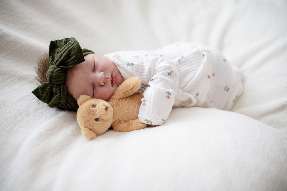 Newborn baby sleeping peacefully in white outfit with green bow headband, cuddling a small teddy bear on white bedding.