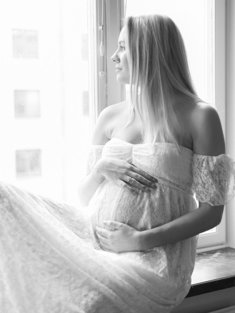 Black and white maternity photo. Person wearing a white dress and is seated by window.