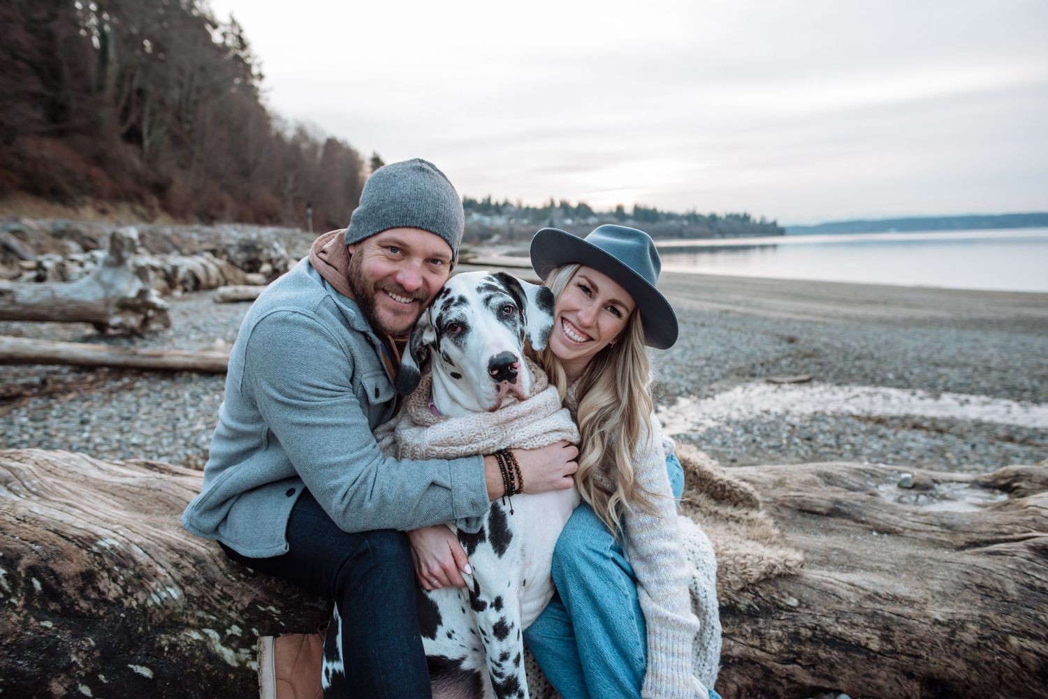 A couple poses with their spotted Dalmatian dog on a rocky beach during an overcast day.