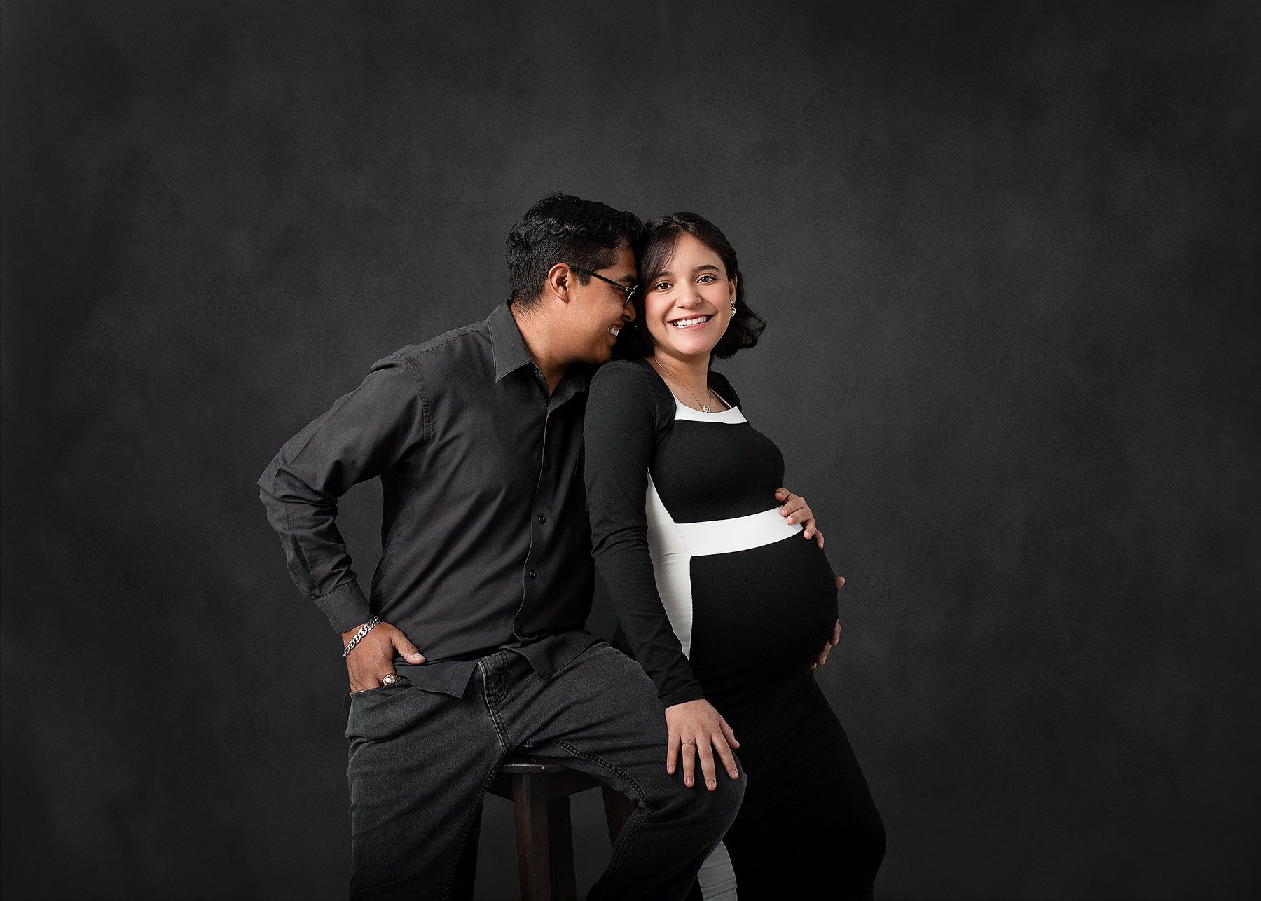 Couple maternity photo shoot, man kissing pregnant woman's cheek, black outfits, studio dark gray background.