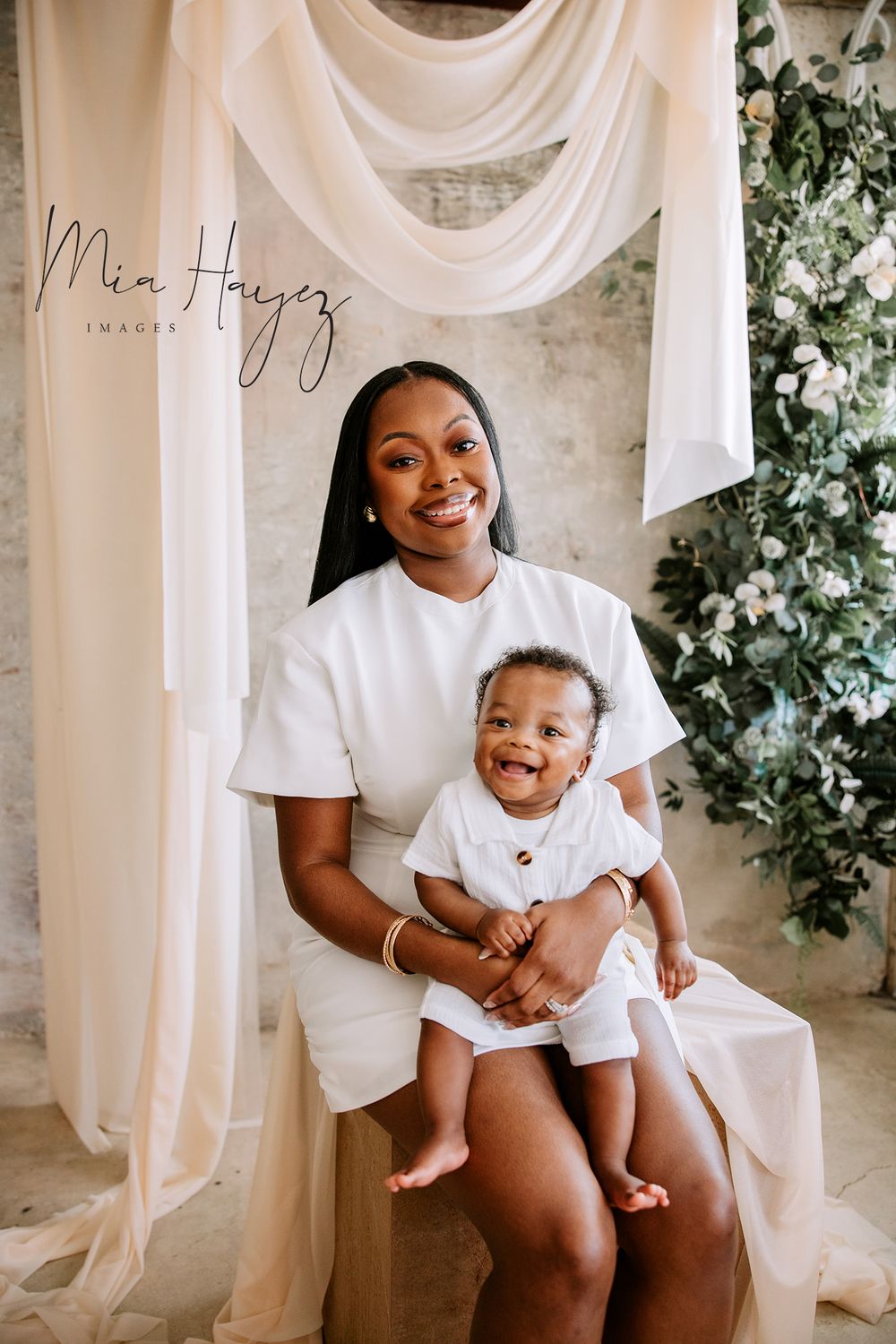 Mother and baby in matching white outfits share tender moments against elegant draped backdrop.
