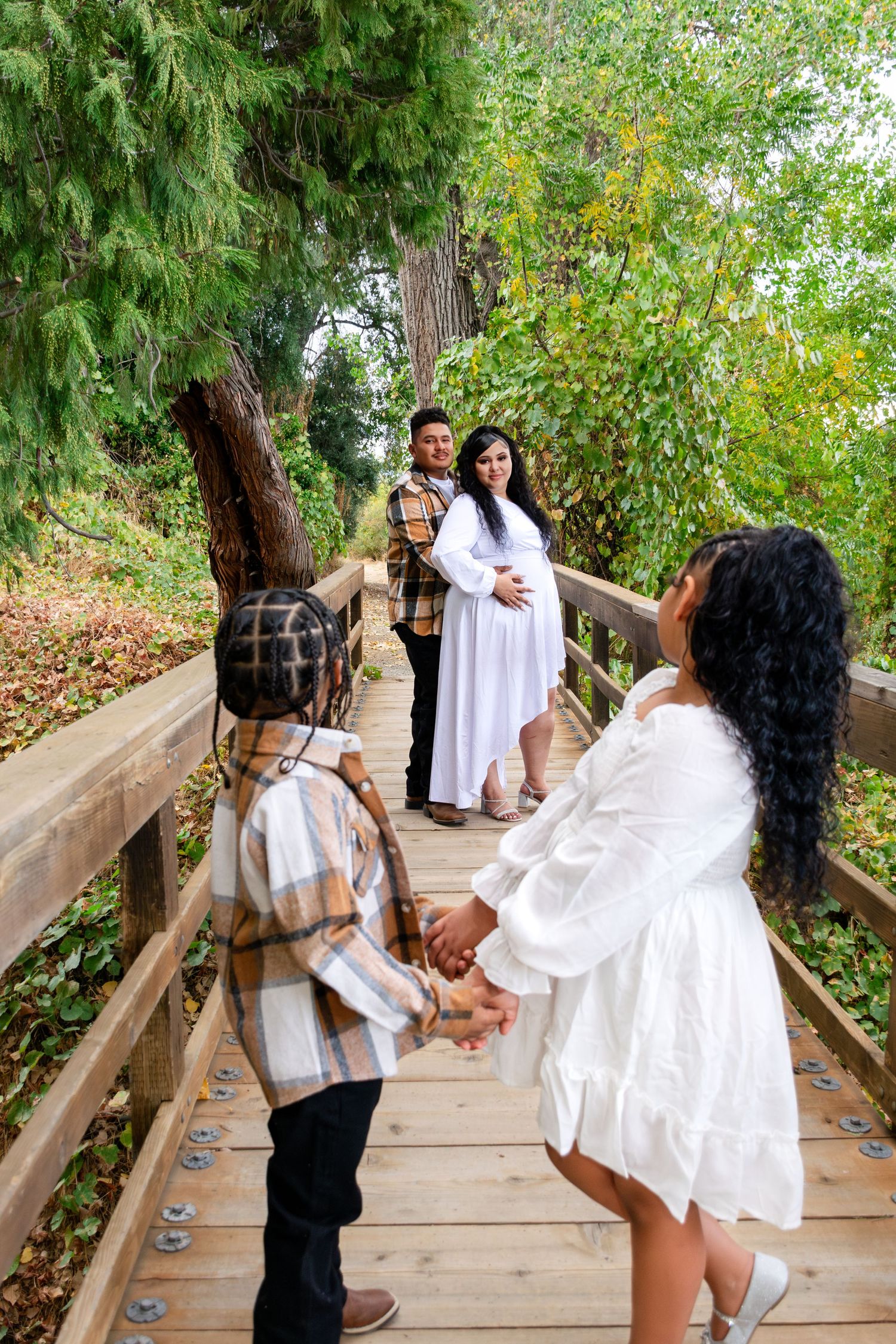 A group wearing coordinated white outfits poses on a wooden bridge surrounded by lush greenery.