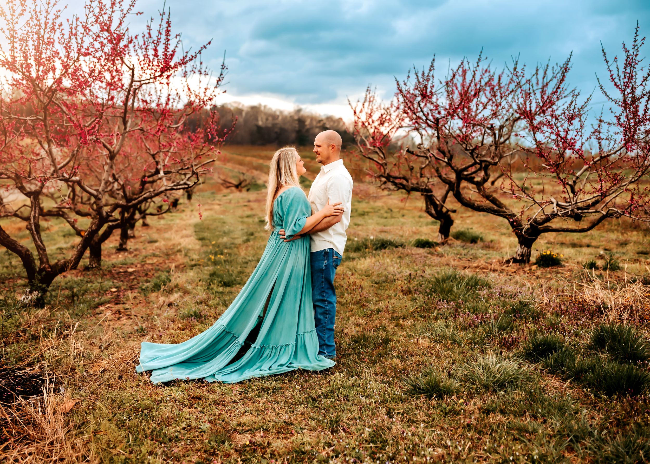 A romantic moment captured in a peach orchard with blooming trees and a woman wearing a flowing turquoise gown.