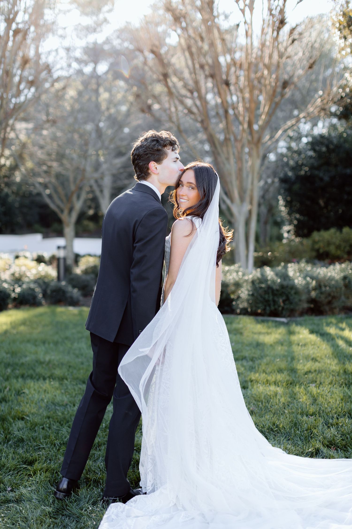 Groom in black tuxedo kisses bride in white gown with long veil in a sunlit garden wedding portrait in Atlanta.