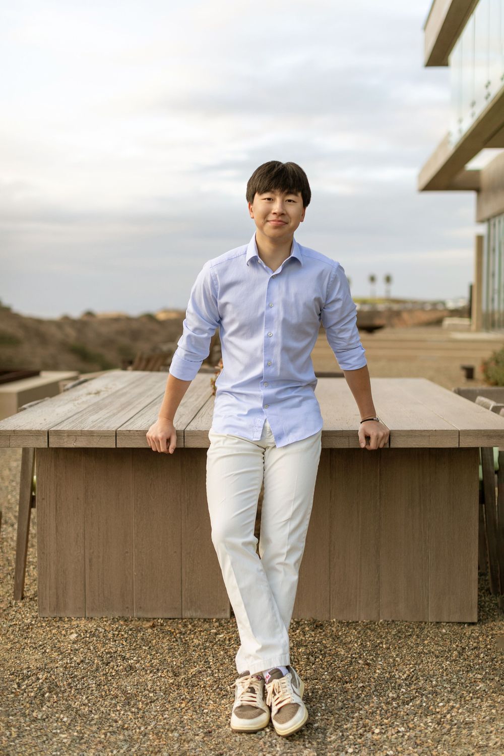 A person in a light blue button-down shirt and white pants leans casually against an outdoor wooden railing at sunset.