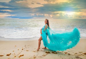 Pregnant woman in flowing teal gown posing on sandy beach with dramatic sunset sky backdrop.