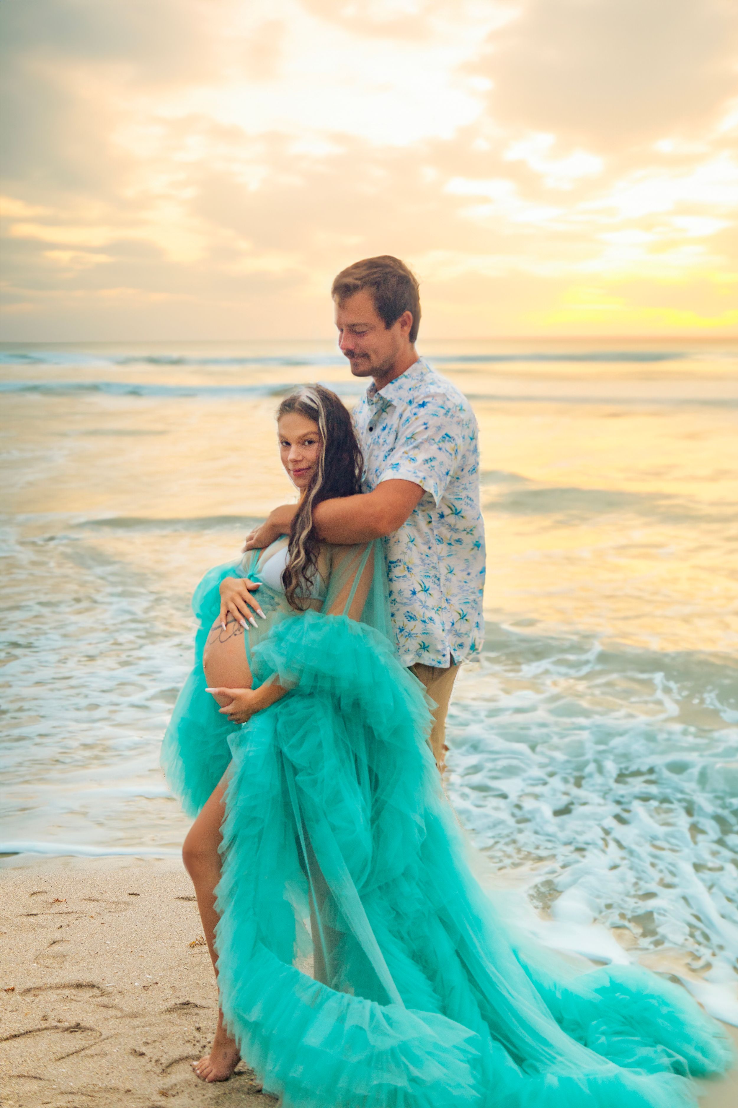 Pregnant woman in flowing teal tulle gown with partner embracing on beach at golden sunset.