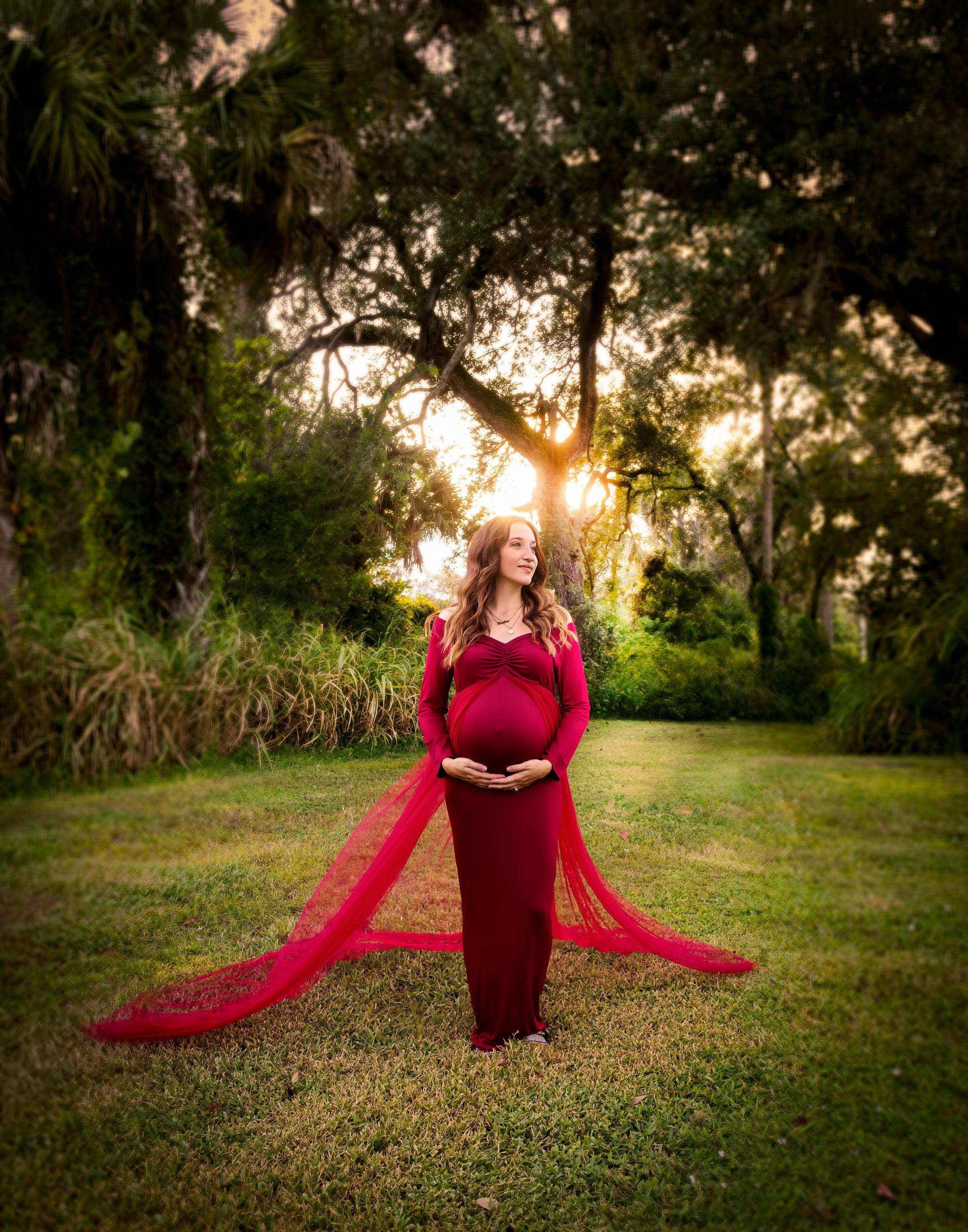 Pregnant woman in red lace maternity gown poses outdoors at golden hour with sunlight filtering through lush green trees.