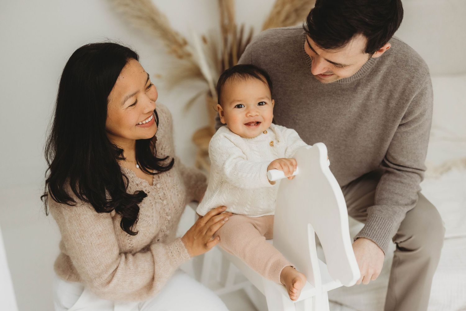 A family in neutral beige and white tones sits together indoors with pampas grass decor in the background.