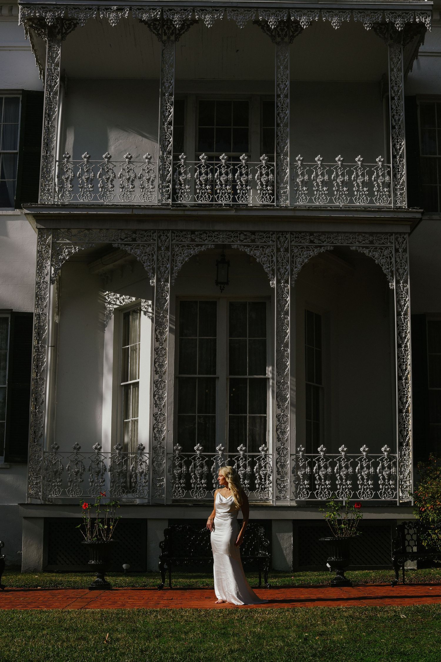 Bride in white dress stands before ornate Victorian mansion with detailed ironwork balcony.