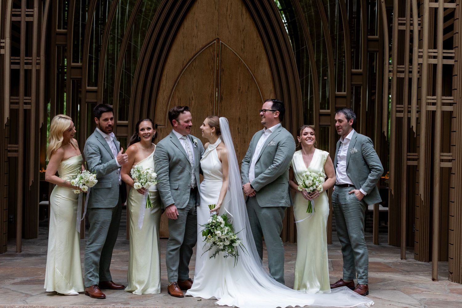 Wedding party wearing light gray suits and pale yellow dresses poses together under gothic arches in a church setting.