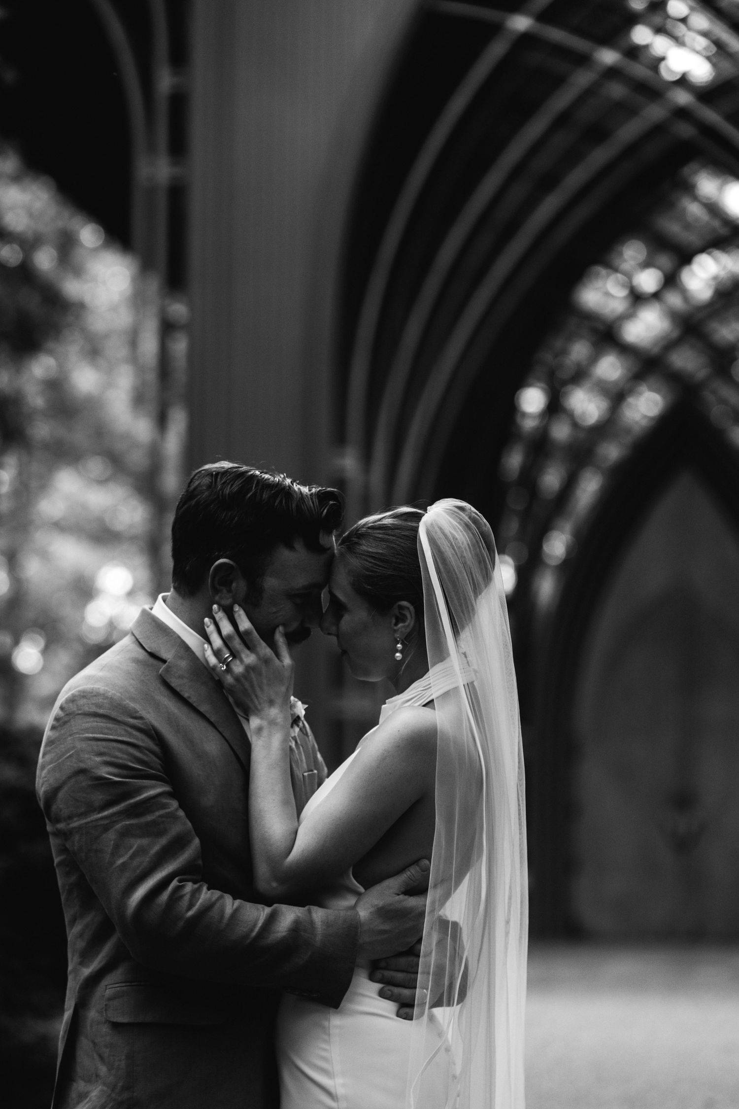 A romantic black and white wedding photograph showing a tender moment between newlyweds beneath an arched cathedral ceiling.